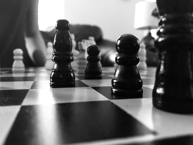 Black chess pieces stand on a checkerboard. A white pawn is visible in the background. The image is in black and white, with a shallow depth of field that blurs the background.