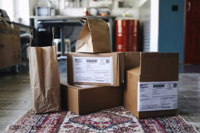 Stacked cardboard boxes and paper bags sit on a patterned rug. Shipping labels are visible on the boxes. A blue door and red canisters are in the blurred background.