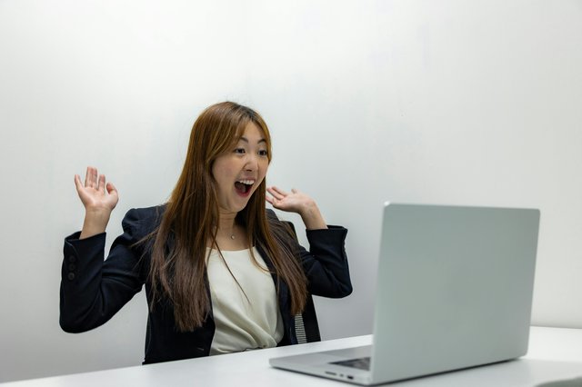 A young woman is seated at a desk, looking directly at the laptop in front of her with a look of excitement and her hands raised in the air. Photo by Resume Genius: https://www.pexels.com/photo/woman-celebrating-in-front-of-a-laptop-in-an-office-18848932/