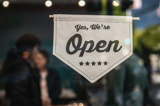 A white banner with black lettering reads "Yes, we're Open." The banner is displayed in a window, with a person's back visible through the glass. String lights hang in the background.